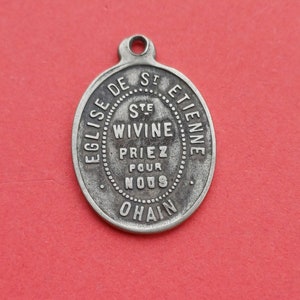 Op de afbeelding: Ovale, zilverkleurige hanger met een lus aan de bovenkant. De hanger heeft gegraveerde Franse tekst, waaronder "EGLISE DE ST ETIENNE" en "STE WIVINE PRIEZ POUR NOUS OHAIN."