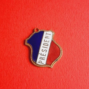May include: A gold-colored metal pin with a blue, white, and red enamel design. The pin features the word "PRESIDENT" in white lettering.