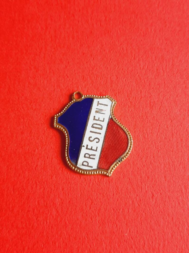May include: A gold-colored metal pin with a blue, white, and red enamel design. The pin features the word "PRESIDENT" in white lettering.