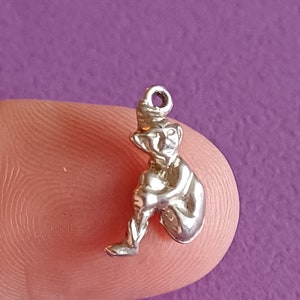 Puede incluir: Un amuleto de plata con forma de elfo sentado. El elfo lleva un gorro puntiagudo y tiene una barba larga.