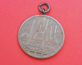 Könnte beinhalten: Runder, bronzefarbener Anhänger mit einem detaillierten Relief eines Gebäudes mit mehreren Türmen und Kuppeln. Der Anhänger hat oben eine kleine Schlaufe zum Befestigen an einer Kette. Der Hintergrund ist einfarbig rot.