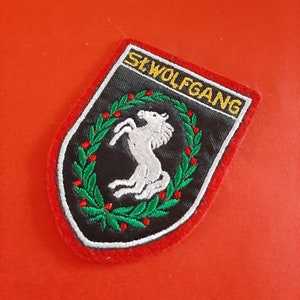 Op de afbeelding: Een zwart en rood geborduurde patch met een wit paard in een groene krans. De tekst "St. Wolfgang" is in geel geborduurd op de patch.