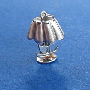 Op de afbeelding: Een zilverkleurige bedel in de vorm van een lamp met een lampenkap en een bootontwerp. De bedel staat op een blauwe achtergrond. De lamp heeft een gedetailleerd ontwerp.