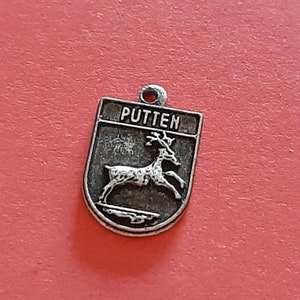 May include: A silver charm with a coat of arms featuring a deer and the word "PUTTEN" on a red background.