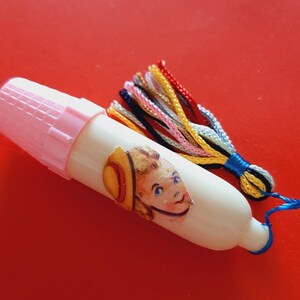 Pu&ograve; includere: Un ditale di plastica bianca con un tappo rosa e un fiocco colorato. Il ditale ha un adesivo di un bambino con un cappello.