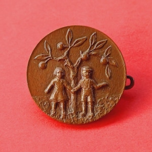 Op de afbeelding: Ronde bronzen speld met een bas-reliëf van twee kinderen die elkaars hand vasthouden onder een fruitboom. De speld heeft een eenvoudige sluiting aan de achterkant en staat tegen een rode achtergrond. Het ontwerp roept nostalgie op.