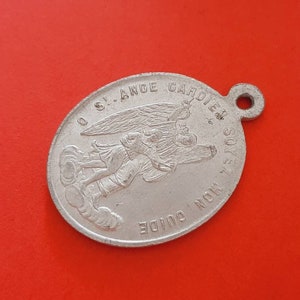 Puede incluir: Un amuleto de metal plateado con una imagen de un ángel sosteniendo a un niño. El amuleto tiene el texto "O.S.I. ANGE GARDIEN DE MON SOIE" y "MON GUIDE" alrededor del borde.