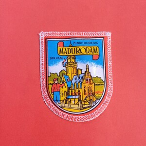 Op de afbeelding: Een Madurom-patch met een kleurrijke illustratie van een miniatuurstad, met gebouwen, een persoon en de tekst "Madurom Den Haag". De patch heeft een wit gestikte rand op een rode achtergrond.