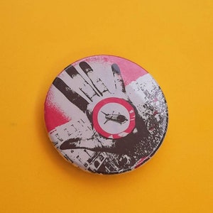 Op de afbeelding: Een ronde knop met een zwart-wit afbeelding van een hand met een rode cirkel en een zwart vliegtuig in de cirkel. De achtergrond is een zwart-wit afbeelding van een stad.