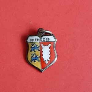 Könnte beinhalten: Ein silberner Anhänger in Form eines Schildes mit dem Wort "NIENDORF" obenauf. Der Schild ist in zwei Abschnitte geteilt, einer gelb mit drei blauen Löwen und der andere rot mit einem weißen stilisierten Berg.