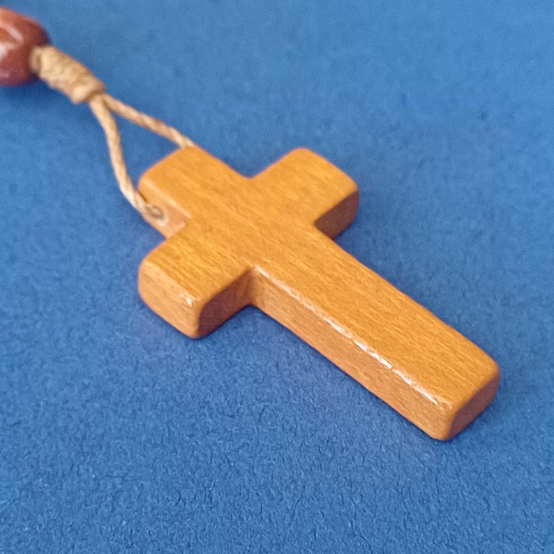 Pu&ograve; includere: Un ciondolo a forma di croce in legno con finitura marrone chiaro, attaccato a un cordino beige e a una serie di perline marroni. La croce &egrave; posta su uno sfondo blu.
