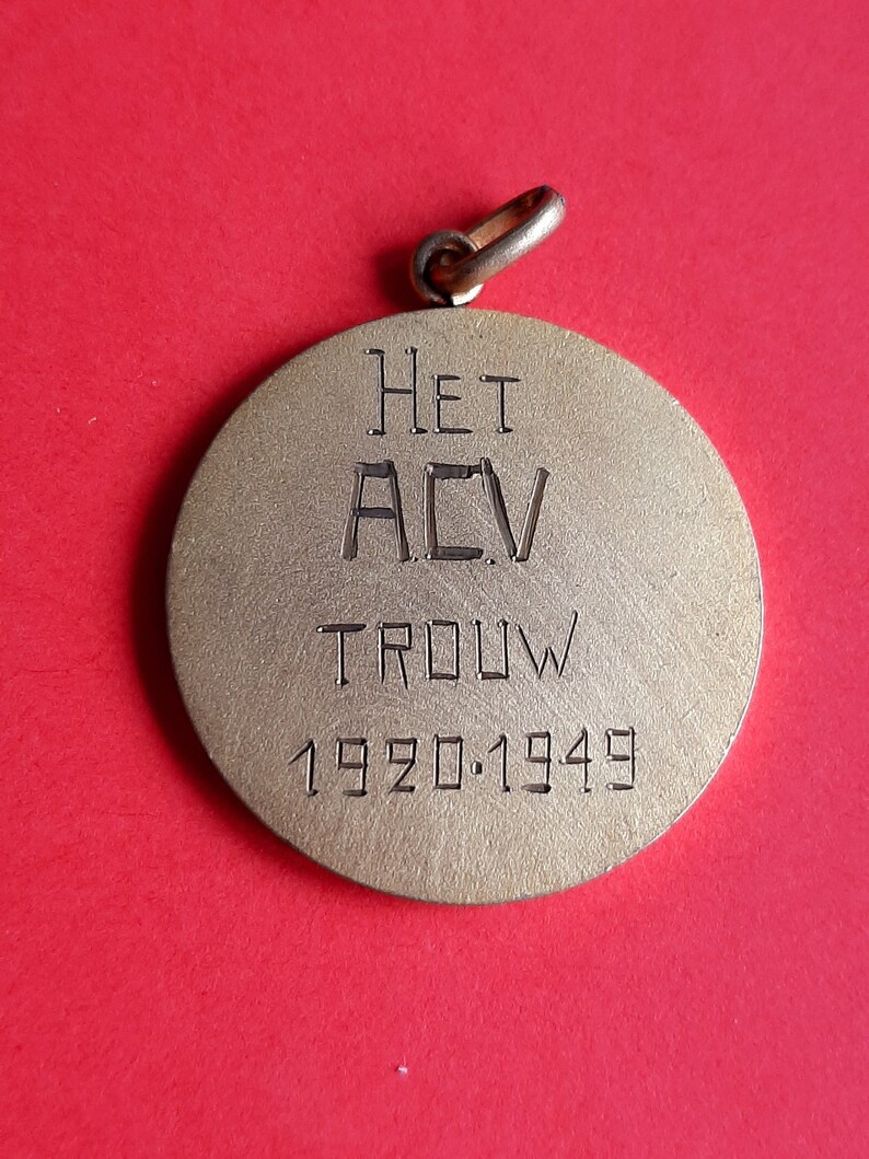May include: A gold-colored metal pendant with the inscription "HET ACV TROUW 1920.1949".