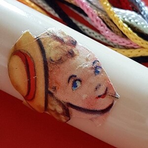Pu&ograve; includere: Un'bobina di plastica bianca con un adesivo colorato di una ragazza che indossa un cappello di paglia. L'adesivo &egrave; leggermente strappato. La bobina &egrave; circondata da filo da ricamo colorato.