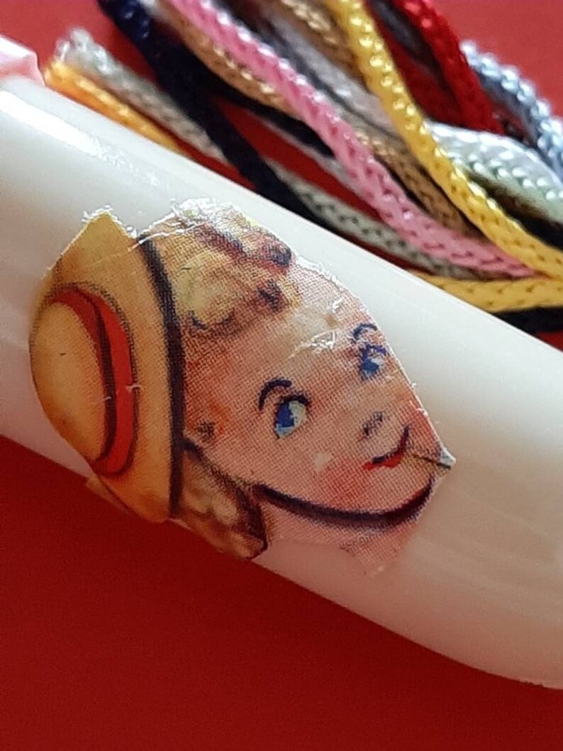 Pu&ograve; includere: Un'bobina di plastica bianca con un adesivo colorato di una ragazza che indossa un cappello di paglia. L'adesivo &egrave; leggermente strappato. La bobina &egrave; circondata da filo da ricamo colorato.