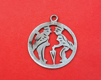 May include: A silver charm with a cutout design of two figures in a circle. The figures are wearing hats and appear to be walking.