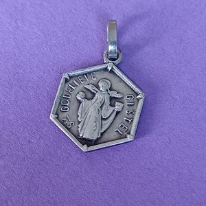 Può includere: Un ciondolo esagonale in argento con un'immagine religiosa di una persona che tiene una croce. Il ciondolo ha le parole "GOD BELIEVE" e "GISTEL" incise.