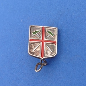 May include: A silver-toned shield-shaped pin with a red cross design. The pin features green and silver details, including two pickaxes. The pin has a small loop at the bottom.