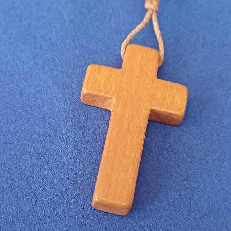 Pu&ograve; includere: Un ciondolo a forma di croce in legno marrone chiaro, appeso a un cordino in fibra naturale. La croce ha un design semplice e classico, con una superficie liscia e bordi arrotondati. Lo sfondo &egrave; blu strutturato.