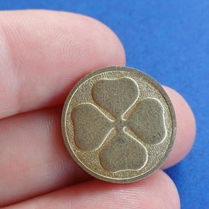 Könnte beinhalten: Eine goldfarbene, kreisförmige Münze oder ein Token mit einem vierblättrigen Kleeblatt-Design. Das Kleeblatt ist in einem strukturierten Kreis geprägt, wodurch ein erhabenes Relief entsteht. Die Münze wird vor einem blauen Hintergrund gehalten.