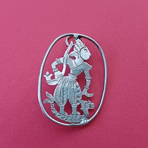 Op de afbeelding: Zilverkleurige hanger met een gedetailleerd uitgesneden ontwerp van een figuur in traditionele kledij, geplaatst in een ovale lijst. De figuur heeft ingewikkelde patronen en staat op een rode achtergrond.