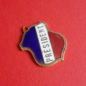 May include: A gold-colored metal pendant shaped like a shield with a blue, white, and red enamel design. The word "PRESIDENT" is written in white on the pendant.