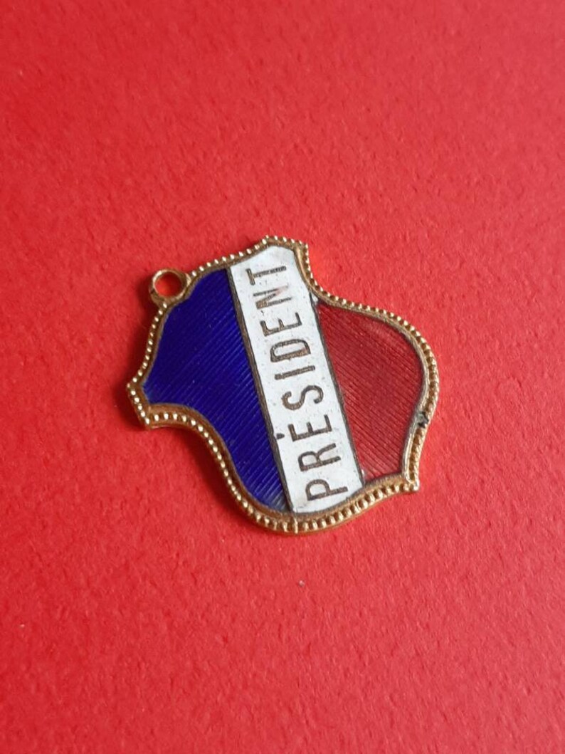 May include: A gold-colored metal pendant shaped like a shield with a blue, white, and red enamel design. The word "PRESIDENT" is written in white on the pendant.