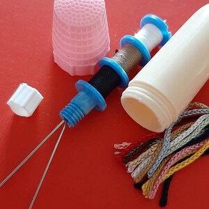 Pu&ograve; includere: Un ditale rosa, un contenitore di plastica bianco con un rocchetto di filo blu e bianco, un rocchetto di filo blu e bianco con filo nero, due aghi argentati e un pacchetto di filo da ricamo colorato.
