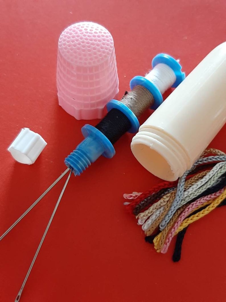Pu&ograve; includere: Un ditale rosa, un contenitore di plastica bianco con un rocchetto di filo blu e bianco, un rocchetto di filo blu e bianco con filo nero, due aghi argentati e un pacchetto di filo da ricamo colorato.