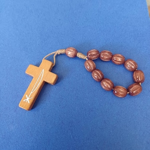 Pu&ograve; includere: Un rosario in legno con perline marroni e una croce. La croce &egrave; marrone chiaro con un disegno bianco. Le perline sono ovali e collegate da un cordoncino intrecciato. Lo sfondo &egrave; blu.