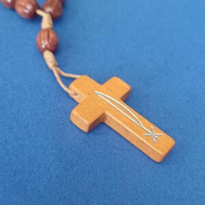 Pu&ograve; includere: Rosario in legno con perline marroni e un ciondolo a forma di croce marrone chiaro. La croce presenta un design intarsiato argentato. Il rosario &egrave; esposto su uno sfondo blu.