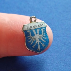 Op de afbeelding: Een kleine, blauw en witte emaille bedel in de vorm van een schild. De bedel toont het woord "ARNHEM" bovenaan en een gestileerd wit adelaarsontwerp. De bedel heeft een zilverkleurige metalen rand en een kleine lus voor bevestiging.