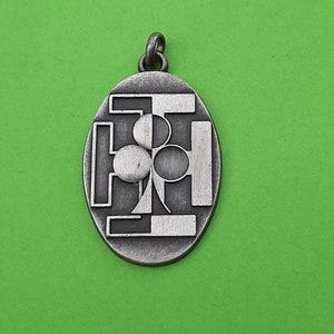 Puede incluir: Un colgante plateado con un diseño de trébol dentro de un marco geométrico. El colgante tiene forma ovalada y tiene un bucle en la parte superior para sujetar una cadena.