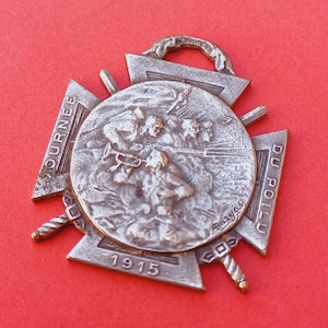 May include: A silver metal medal with a cross design. The center of the medal features a relief sculpture of soldiers in battle. The medal is inscribed with the text "JOURNEE DU POILU 1915".