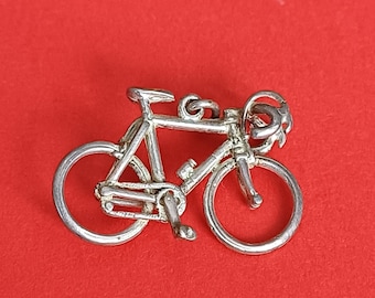 May include: A silver-toned metal charm shaped like a bicycle. The charm features a frame, two circular wheels, handlebars, a seat, and pedals. A small circular jump ring is attached to the top of the frame for hanging or connecting to a chain.