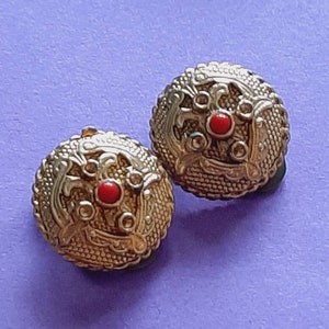May include: A pair of gold-tone clip-on earrings with a red stone in the center of each earring. The earrings have a detailed design with intricate patterns.