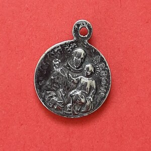 May include: Silver charm featuring a depiction of Saint Anthony of Padua holding a baby Jesus. The charm has a loop at the top for attaching to a necklace or bracelet.