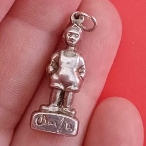 May include: A small, silver-colored charm in the shape of a person wearing overalls, with hands in pockets. The charm is standing on a rectangular base with the word "Barbie" engraved on it. The charm has a loop at the top for attaching to a bracelet or necklace.