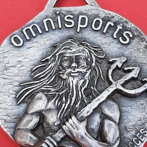 May include: A silver-toned metal pendant with a raised image of a bearded man holding a trident. The pendant has the word "omnisports" inscribed around the top edge and the word "success" inscribed at the bottom.