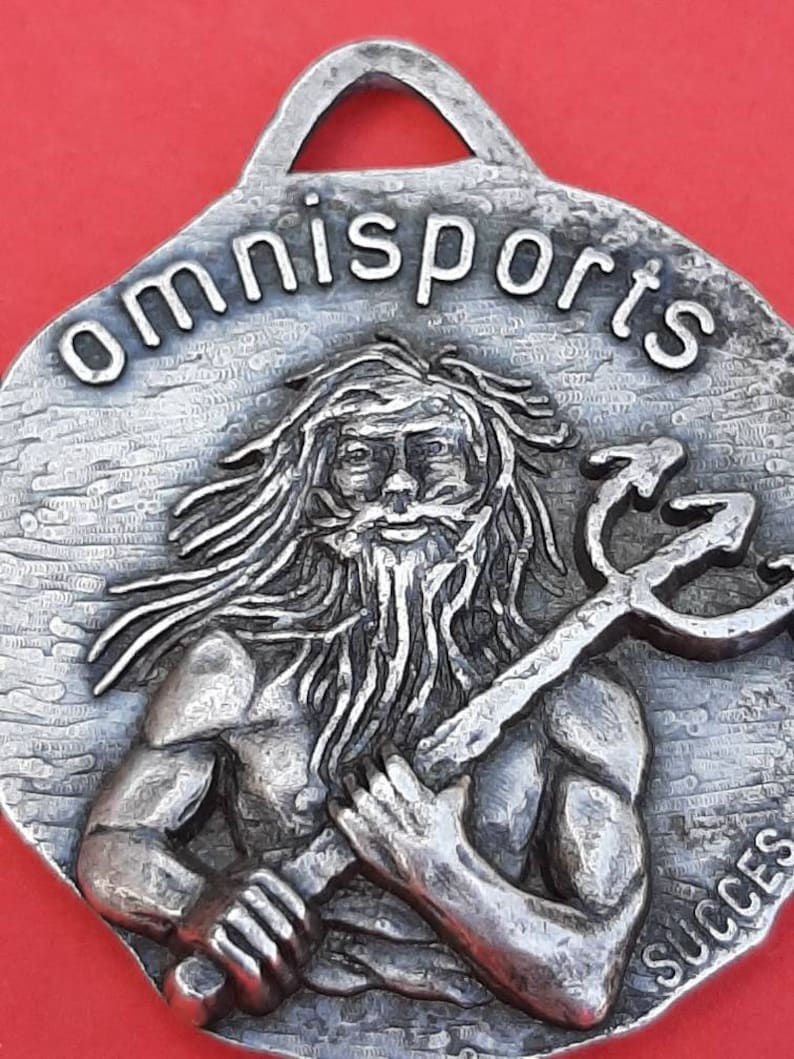 May include: A silver-toned metal pendant with a raised image of a bearded man holding a trident. The pendant has the word "omnisports" inscribed around the top edge and the word "success" inscribed at the bottom.