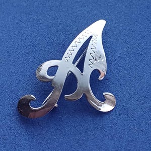 Może przedstawiać: Srebrna broszka w kształcie litery "A". Broszka ma dekoracyjny, kursywny wzór z zygzakowatym wzorem na górze i na dole litery. Broszka jest umieszczona na niebieskim tle.
