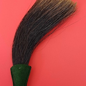 Peut inclure: Une brosse incurvée avec des cheveux foncés et une poignée en feutre vert. La brosse présente un mélange de poils noirs et bruns, avec des pointes plus claires. La poignée est vert foncé, sur fond rouge uni.