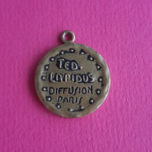 Könnte beinhalten: Ein goldfarbener Metallanhänger mit dem Text "TED. LAPIDUS DIFFUSION PARIS" in schwarzer Schrift.