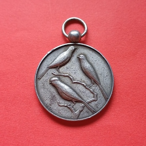 May include: A circular, silver-coloured pendant depicting three birds on a branch. The birds are in a circular frame with a loop at the top for hanging. The background is a solid red.