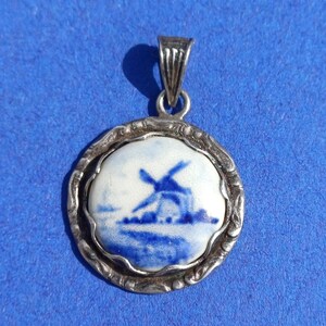 Pode incluir: Pingente redondo com moldura e argola prateadas. O pingente apresenta uma cena de moinho de vento pintada em azul e branco. A moldura tem uma borda decorativa e texturizada. O pingente é provavelmente feito de cerâmica ou porcelana.