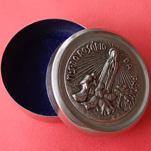 Op de afbeelding: Open zilverkleurige ronde doos met een blauw fluwelen interieur. Het deksel heeft een gedetailleerd reliëf van religieuze figuren en het woord "ROSÁRIO". De doos staat tegen een rode achtergrond.