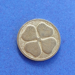 Könnte beinhalten: Eine goldfarbene Münze mit einem Kleeblatt-Design. Die Münze hat einen strukturierten Rand und befindet sich auf einem blauen Hintergrund. Die Blätter des Kleeblatts sind herzförmig.