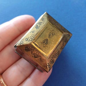 Op de afbeelding: Een kleine, rechthoekige, antieke messing doos met ingewikkelde bloemen- en bladgravures. De doos heeft een scharnierend deksel en een aangetaste, verouderde uitstraling, wat suggereert dat hij vintage is. De doos wordt in een hand gehouden tegen een blauwe achtergrond.