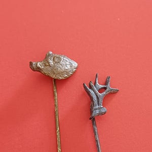 Puede incluir: Dos alfileres de sombrero antiguos. Uno presenta una cabeza de jabalí detallada en tono plateado, y el otro tiene un diseño de cornamenta estilizado. Ambos alfileres tienen varillas largas y delgadas. El fondo es rojo liso.