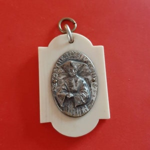 May include: A white oval pendant with a silver image of a person in a robe. The pendant is inscribed with the text "Saint Martin Lequeux".