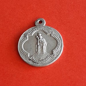May include: Silver pendant featuring a religious image of the Virgin Mary holding a baby Jesus. The pendant has a detailed design with a scalloped edge.